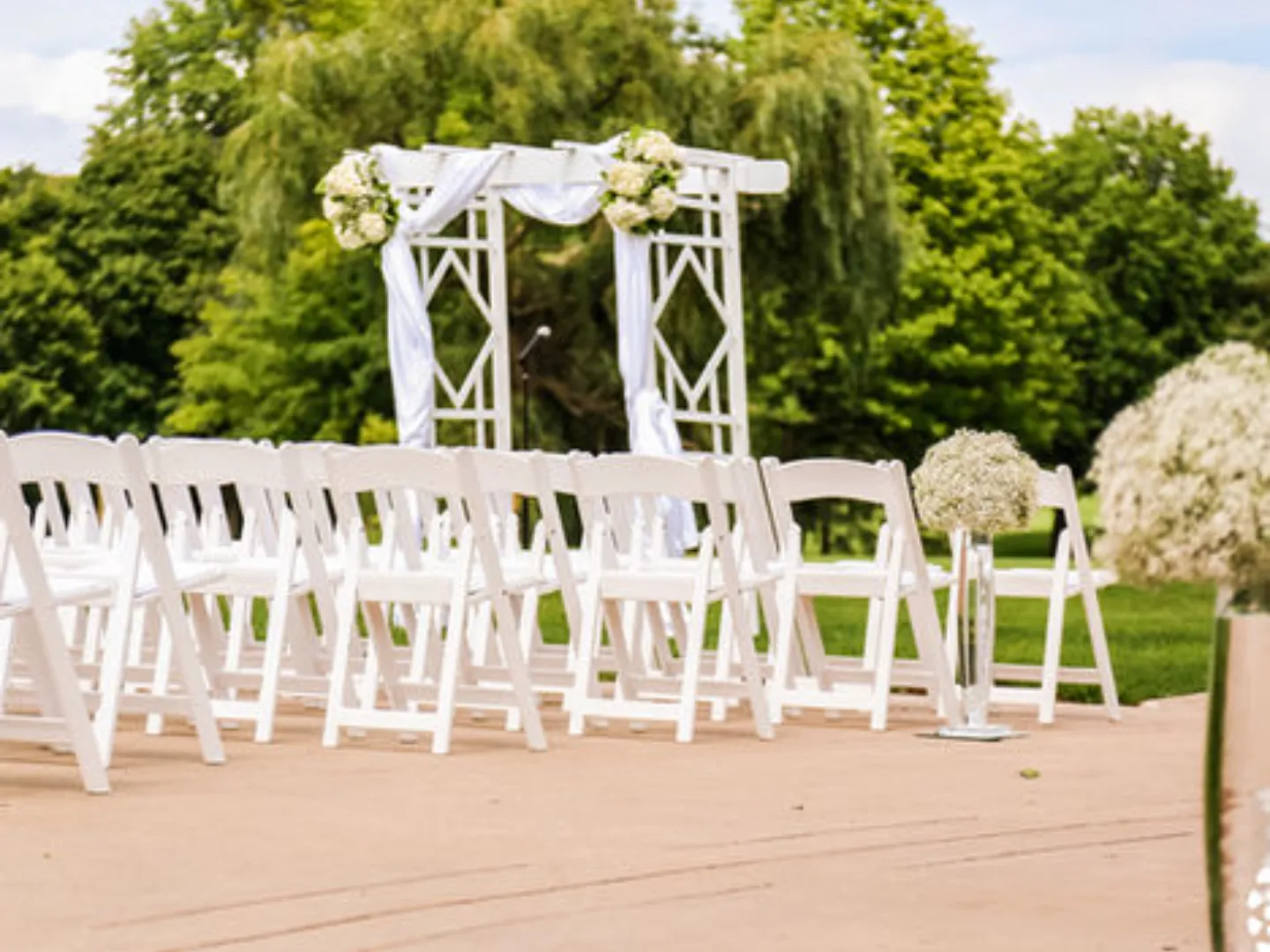 A white wooden ceremony arch dressed with flowers in front of white chairs and green trees