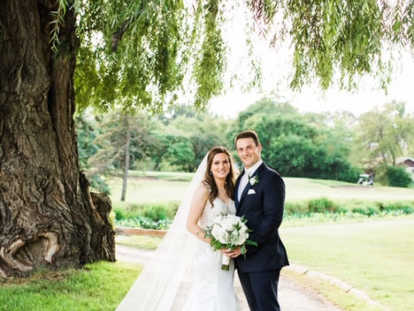 A bride and groom pose beneath a large willow tree beside the course
