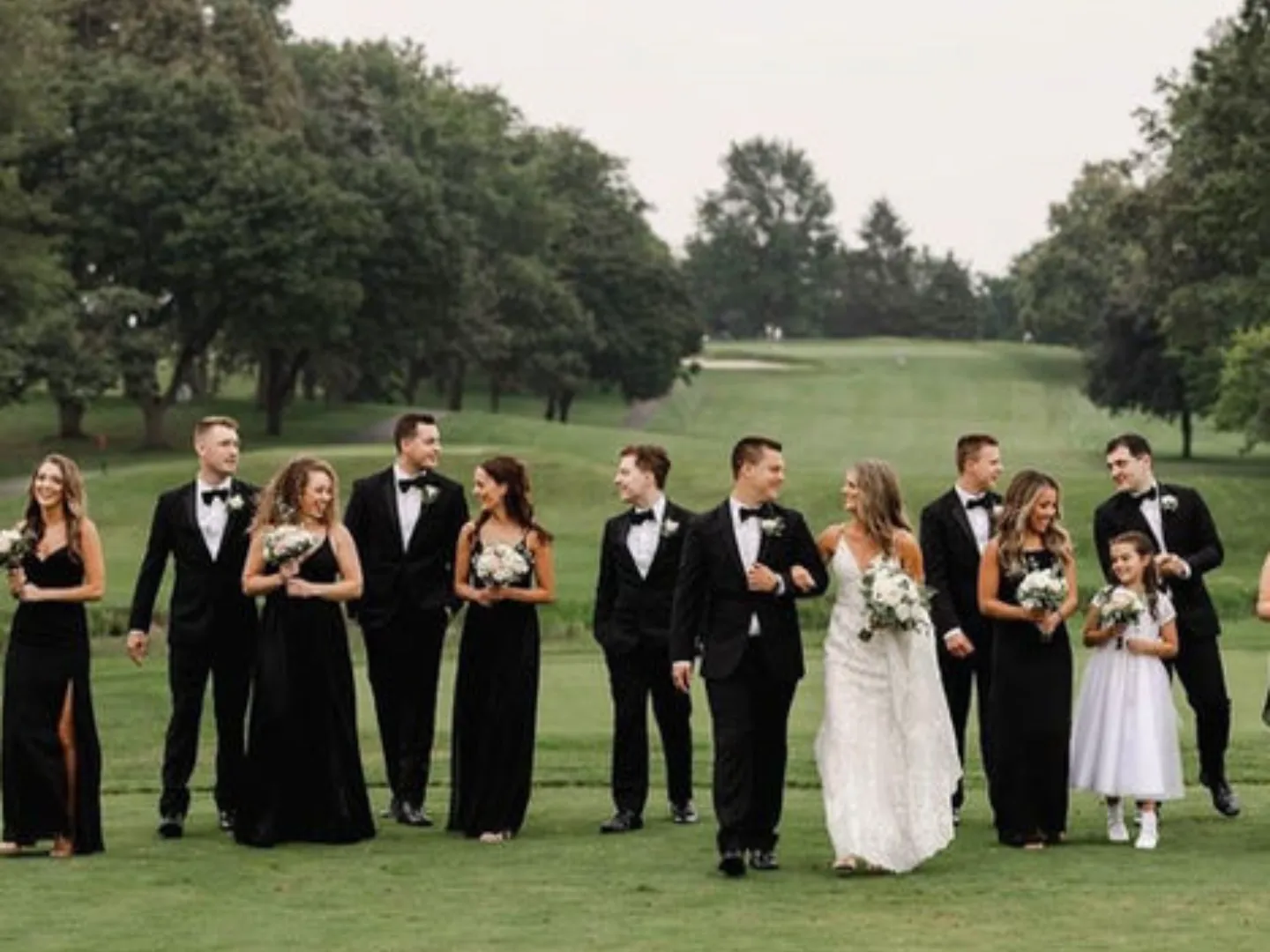 A full wedding party walks together down a fairway lined with trees