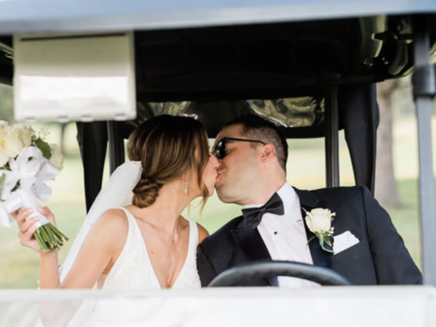 A bride and groom kiss in the front seat of a golf cart