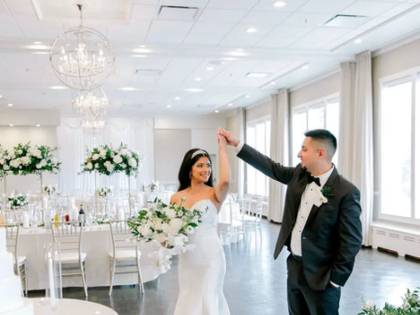 A groom twirls his bride in the bright white Grand Ballroom with tall floral centerpieces and chandeliers