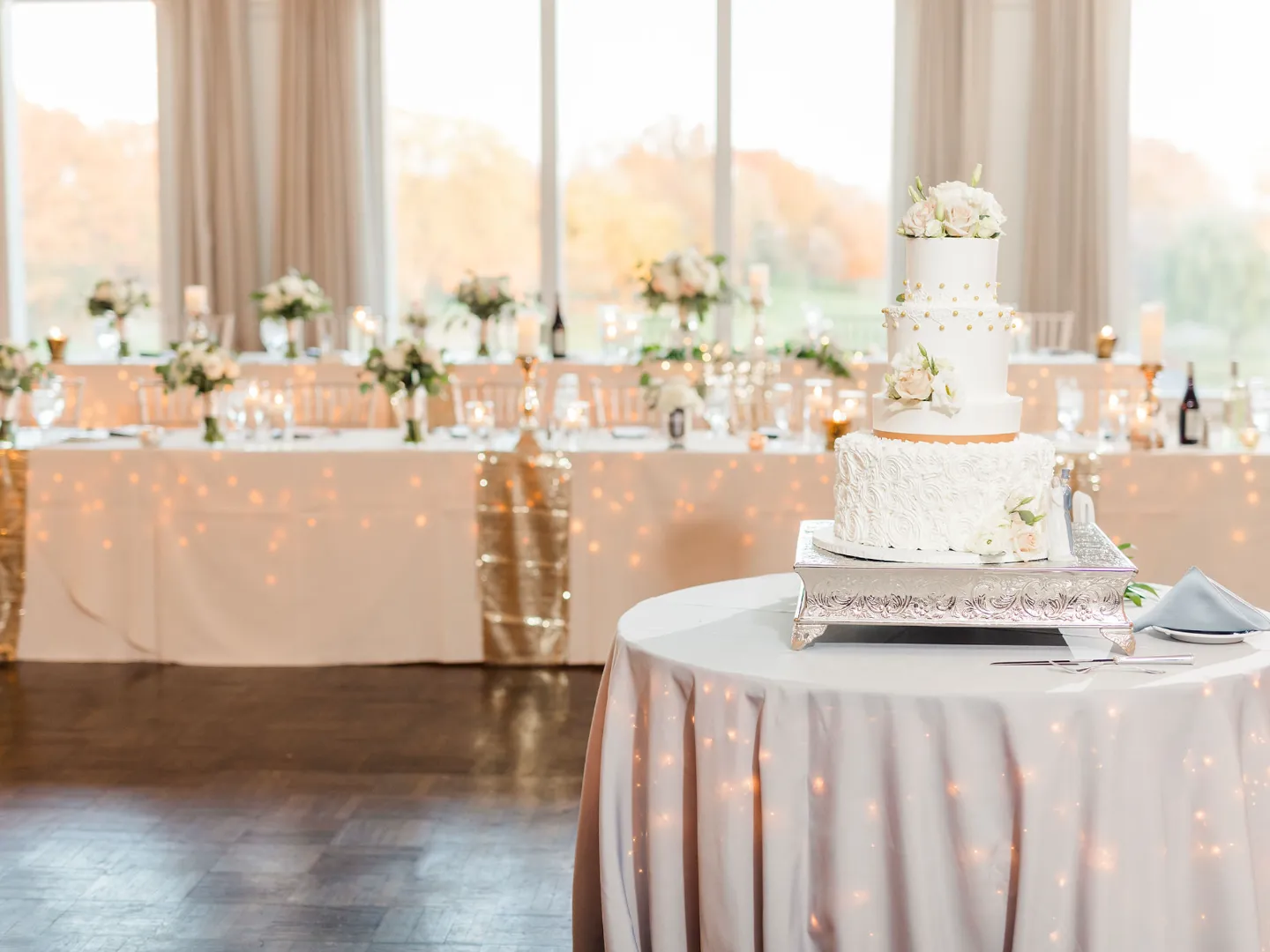 A tiered white wedding cake in front of a head table lined with flowers and candles