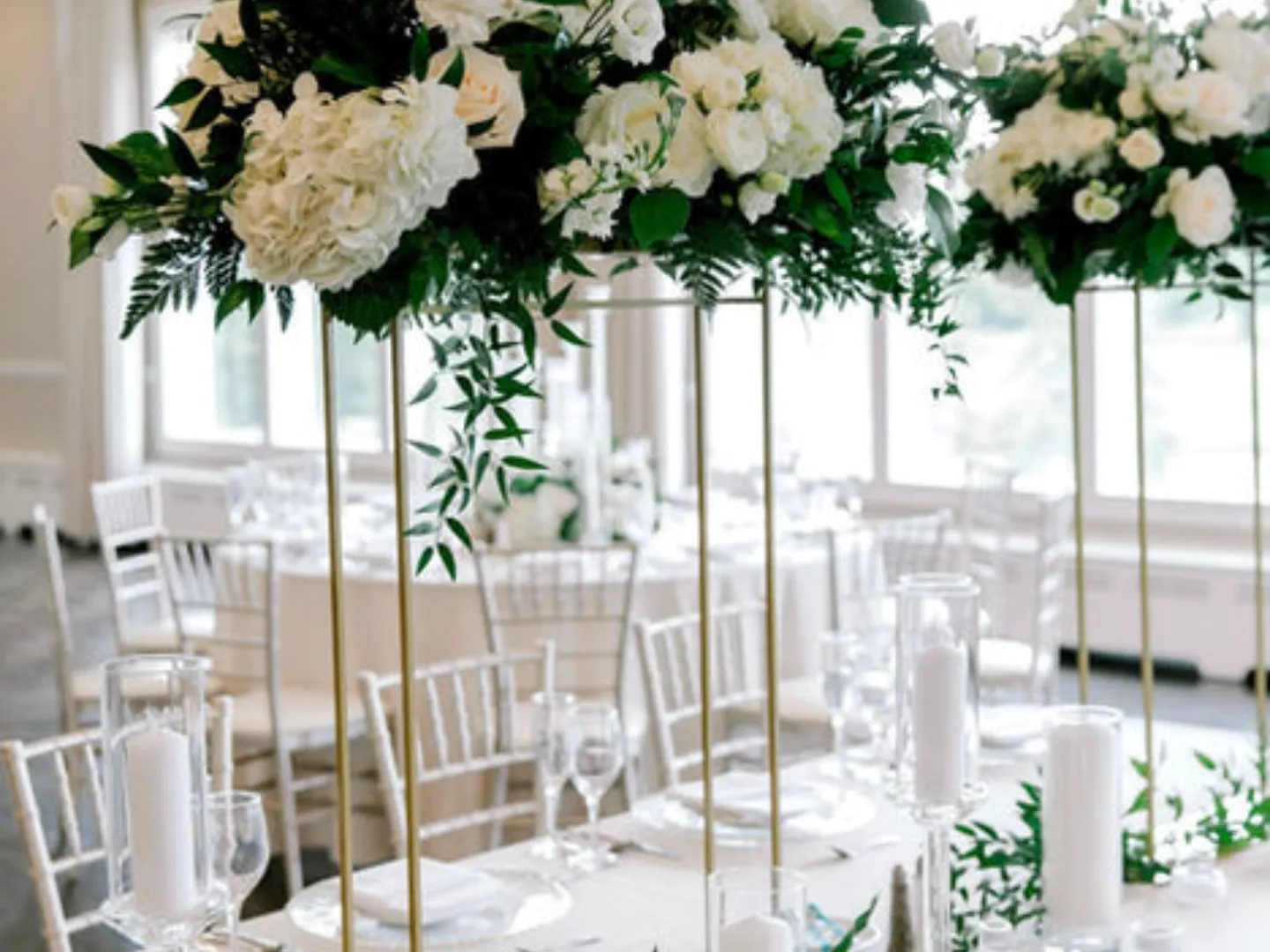 Reception tables with white linens, candles and tall white floral arrangements by the windows