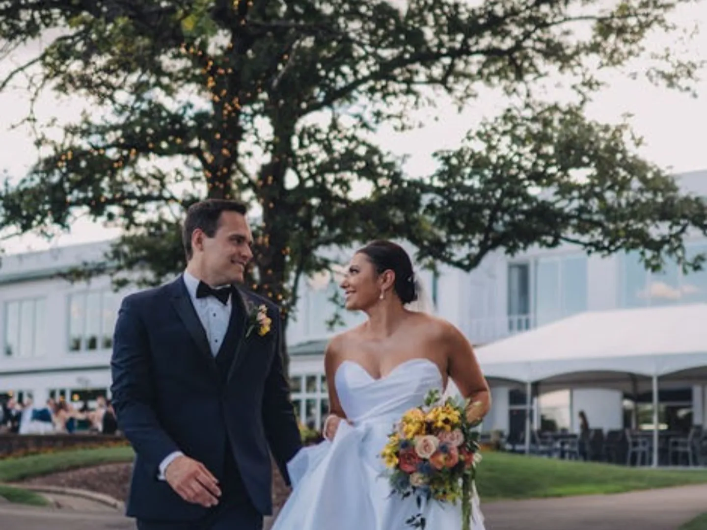 A groom in a navy tuxedo and a bride holding a bright bouquet laugh together outside the clubhouse