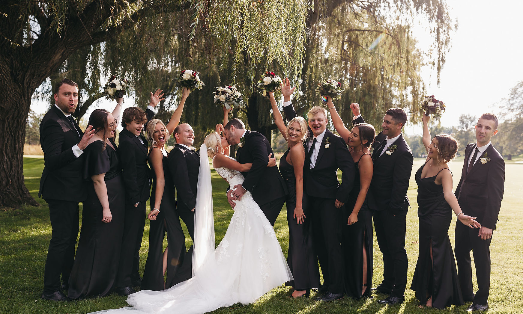 A cheering wedding party in black gowns and tuxedos under a willow tree